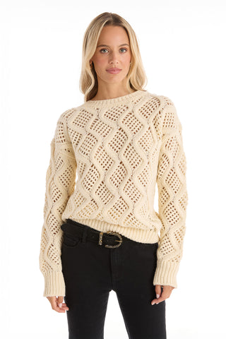 A woman with long blonde hair wears the Sonja Wave Sequin Sweater - Pearl, a cream knit with wavy patterns and sequins, paired with black jeans, standing against a plain white background.