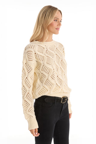 A woman with long blonde hair wears the Sonja Wave Sequin Sweater - Pearl, featuring a textured wave pattern, paired with black jeans and a belt. She stands against a white background, facing slightly right and smiling gently.