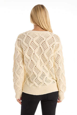 A woman with long blonde hair is seen from the back wearing the Sonja Wave Sequin Sweater - Pearl, a cream knit with a wavy textured pattern and subtle sequins, paired with sleek black pants.