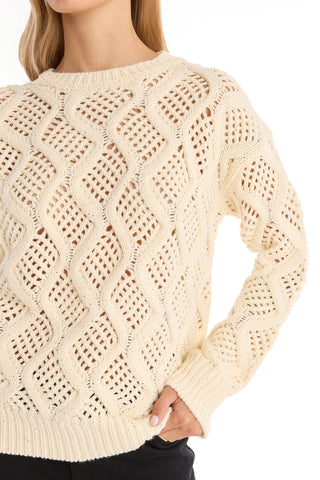 A woman wears the Sonja Wave Sequin Sweater - Pearl, a cream knit with wave patterns, ribbed cuffs, hem, and collar. Her left hand is on her hip as she effortlessly pairs it with black pants.