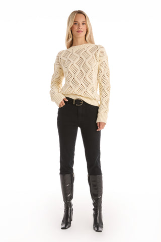 A woman faces forward wearing the Sonja Wave Sequin Sweater - Pearl, which features a knit wave pattern and diamond accents, paired with black pants, a black belt, and knee-high boots against a white background.