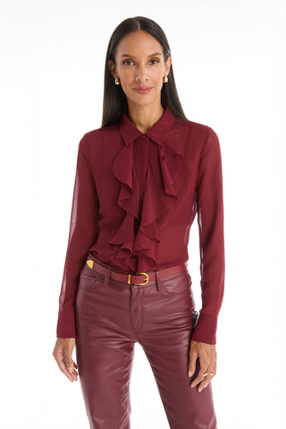 A woman stands against a white background, wearing the Georgia Semi-Sheer Blouse in burgundy and matching leather pants. With long dark hair and gold hoop earrings, she exudes modern elegance in this romantic, ruffle-detailed statement piece.