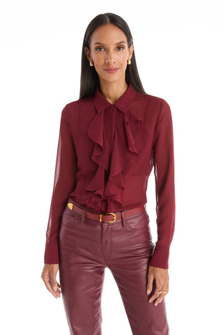A woman faces forward in a burgundy Georgia Semi-Sheer Blouse with ruffle details and matching leather pants. She has long dark hair, gold hoop earrings, and a neutral expression against a white backdrop.