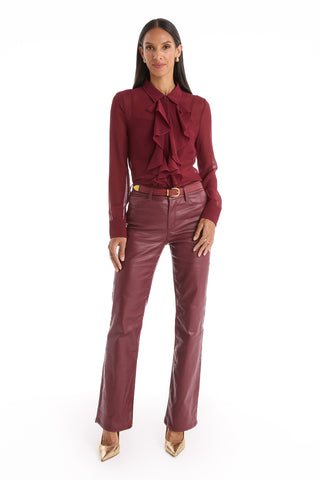 A woman with long dark hair wears a burgundy ruffled blouse and Aria Coated Flare Jean in Merlot with a high-rise fit, gold-buckled belt, and gold pointed-toe heels. She stands facing forward against a plain white background.