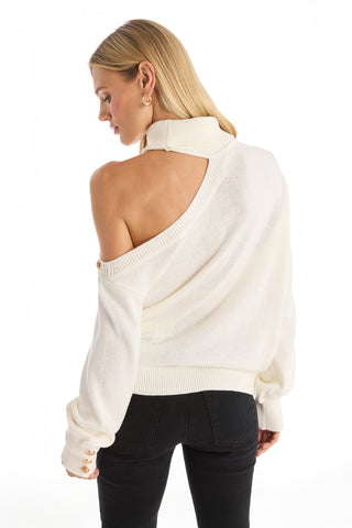 A woman with straight blonde hair models the Bridgette Button Sweater - Pearl, wearing it off one shoulder with black pants and facing away from the camera against a white background, highlighting chic modern style.