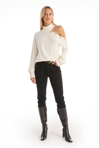 A woman faces forward, wearing the Bridgette Button Sweater - Pearl—an asymmetric white knit with gold button details and one shoulder exposed—paired with black jeans and knee-high boots against a plain white background.