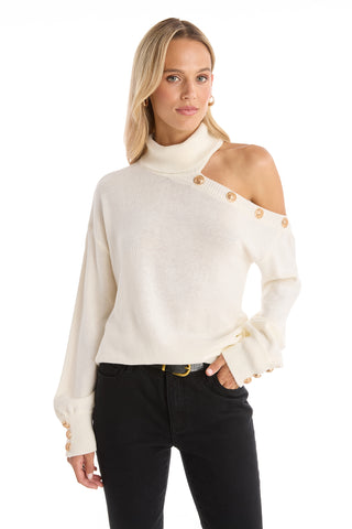 A woman wears the Bridgette Button Sweater - Pearl, an asymmetrical cream knit with gold shoulder and cuff buttons that exposes one shoulder. She pairs it with black pants and a belt, standing against a white background.
