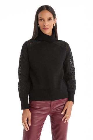 A woman wears the Camille Lace Turtleneck Sweater in black, featuring delicate lace details on the shoulders and sleeves, paired with high-waisted burgundy leather pants, standing against a plain white background.
