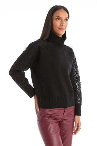 A woman in the Camille Lace Turtleneck Sweater – Black, featuring delicate lace details, stands with one hand on her hip and smiles while looking to the side.
