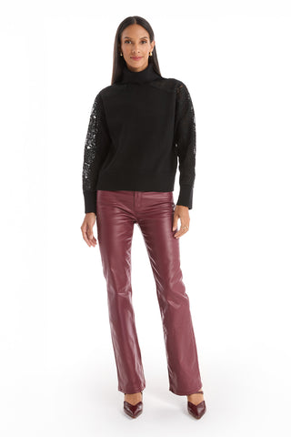 A woman stands against a white background wearing the Camille Lace Turtleneck Sweater in black, detailed sleeves, styled with burgundy leather pants and matching heels.