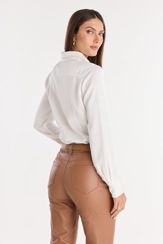 A woman with long brown hair, wearing the Paulina Silk Blouse in white and high-waisted light brown leather pants, stands with her back turned and looks over her shoulder against a white background.