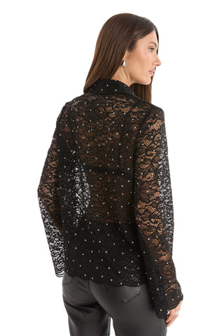 A woman with long brown hair models the Jena Lace Button Up Shirt in black, a sheer floral lace blouse with sparkling details, paired with black leather pants. She is turned to the side, showcasing the shirt’s back against a white background.
