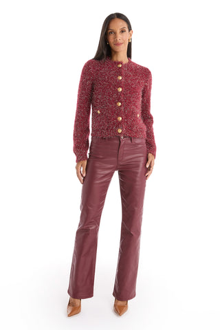 A woman with long dark hair faces forward, wearing the Joana Knit Cardigan - Merlot with gold buttons and padded shoulders, paired with burgundy leather pants and tan heels, standing against a white background.