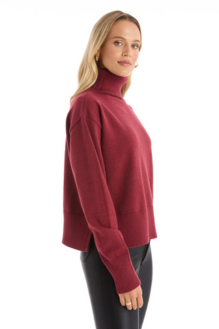 A woman with long blonde hair models the Gia Ultra Soft Sweater - Merlot, an oversized turtleneck pullover, paired with black pants. She stands sideways, looking at the camera against a plain white background.