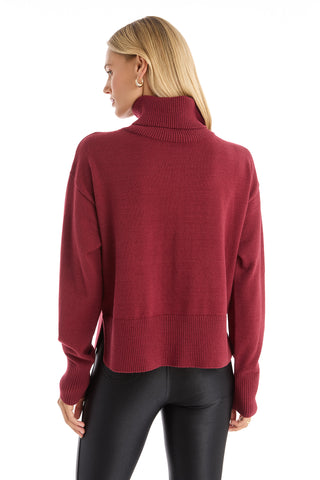 A woman with straight blonde hair is shown from the back, wearing the Gia Ultra Soft Sweater - Merlot, a relaxed tunic with long sleeves and an oversized turtleneck, paired with black shiny pants against a white background.