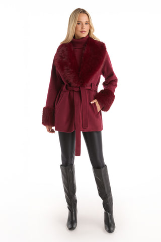A woman wears the Isabel Fur Trim Coat in Merlot, showcasing its timeless silhouette with a belted waist, faux fur collar and cuffs, paired with black leggings and knee-high boots against a white background.