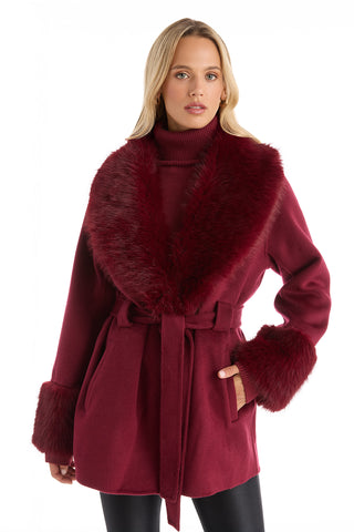 A woman with long blonde hair and a neutral expression models the Isabel Fur Trim Coat - Merlot, featuring a belted waist and fur trim, against a white background, highlighting the classic elegance of this timeless outerwear.