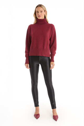 A woman faces forward wearing the Gia Ultra Soft Sweater - Merlot, an oversized turtleneck pullover styled with black shiny leggings and red pointed-toe heels against a white background.