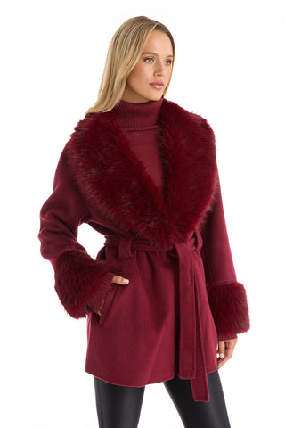 A woman poses against a white background wearing the Isabel Fur Trim Coat in Merlot, a timeless silhouette with a belted waist and faux fur collar and cuffs, styled over a matching turtleneck sweater.