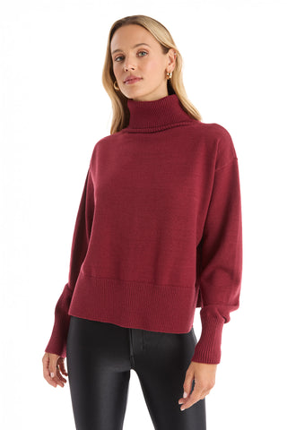 A woman wearing the Gia Ultra Soft Sweater in Merlot, an oversized long-sleeve turtleneck pullover, and black fitted pants stands facing forward against a white background.