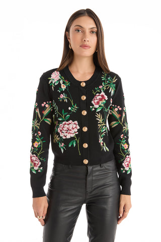 A woman with long straight hair and a neutral expression faces forward, wearing the Alyssa Jacquard Cardigan in black floral with colorful embroidery and gold buttons, paired with black leather pants. The background is white.