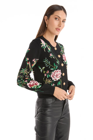Woman in the Alyssa Jacquard Cardigan - Black Floral, featuring gold buttons and padded shoulders, paired with black leather pants and standing against a white background.