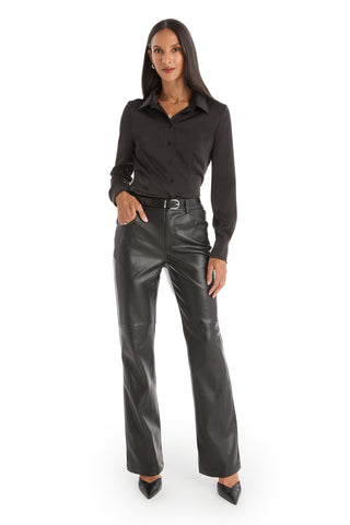 A woman stands confidently wearing the Paulina Silk Blouse - Black, tucked into black high-waisted faux leather pants with a belt and black heels, posed against a plain white background.