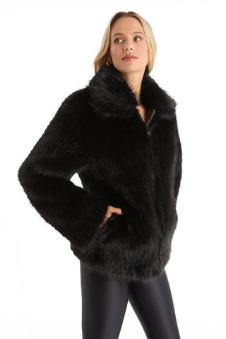 A woman with shoulder-length blonde hair stands against a white background, hands in the pockets of the Dani Fur Coat - Black, showcasing its chic, cruelty-free faux fur style.