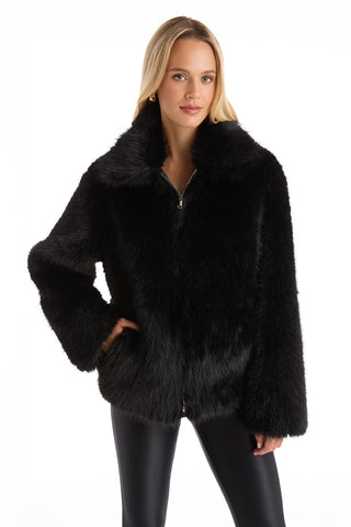 A woman with long blonde hair wears the Dani Fur Coat - Black and black leather pants, standing with one hand in her pocket against a plain white background.