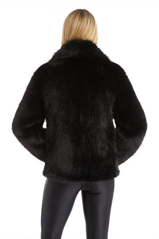 A person with long blonde hair, seen from behind, wears the Dani Fur Coat - Black, a cruelty-free faux fur jacket, along with shiny black pants against a white background.