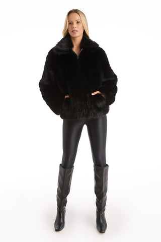 A woman faces forward against a white background, hands in her pockets, wearing the Dani Fur Coat - Black with shiny black leggings and knee-high black boots.