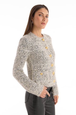 A woman with long brown hair wears the Joana Knit Cardigan - Grey, featuring a textured knit, padded shoulders, and large yellow buttons, paired with black pants. She stands against a plain white background, looking slightly to the side.