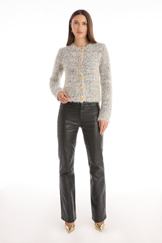 A woman faces forward in the Joana Knit Cardigan - Grey with gold buttons, paired with black leather pants and gold pointed-toe heels, set against a plain white background.