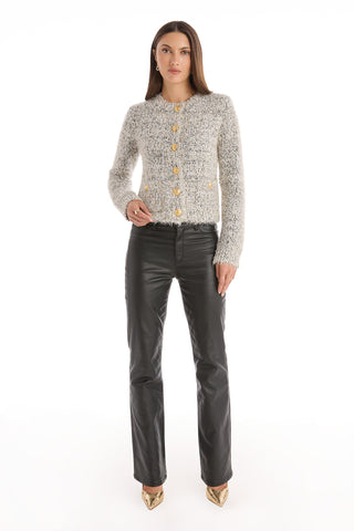 A woman with long straight hair and a neutral expression stands against a white background, wearing the Joana Knit Cardigan - Grey, paired with black leather pants and gold pointed-toe heels.