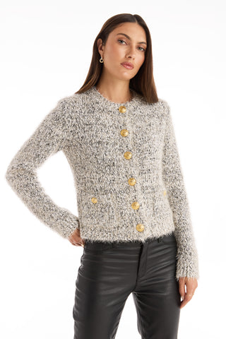 A woman with straight brown hair wears the Joana Knit Cardigan - Grey, featuring padded shoulders and gold buttons, paired with black leather pants. She stands against a white backdrop with one hand on her hip.