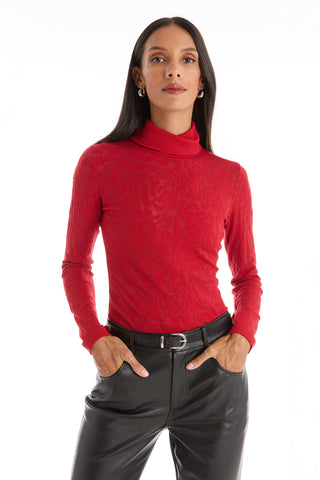 A woman with long dark hair models the Alina Rose Turtleneck Top - Red, featuring a subtle embossed pattern, paired with black leather pants. She stands with her hands in her pockets against a white background.