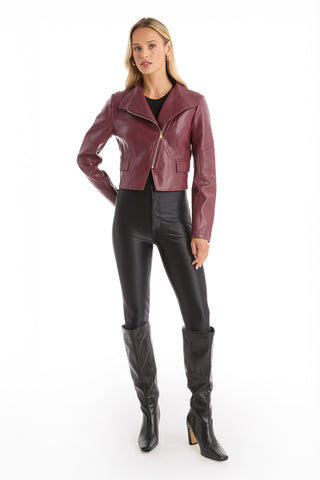 A woman with long blonde hair wears the Katalina Moto Jacket - Merlot, featuring a sleek front zip, over a black top with high-waisted black pants and knee-high boots, posing against a plain white background.