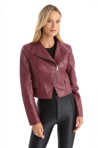 A woman with long blonde hair wears the Katalina Moto Jacket - Merlot, featuring a sleek front zip closure, over a black top and pants, standing against a plain white background.