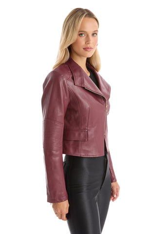 A woman with long blonde hair wears the Katalina Moto Jacket - Merlot, featuring a sleek front zip and asymmetrical silhouette, paired with black pants as she stands slightly to the side against a white background.