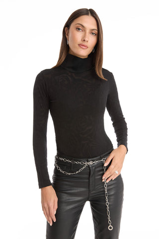 A woman with straight brown hair and hoop earrings stands against a white background wearing the Alina Rose Turtleneck Top - Black, styled with black leather pants and a silver chain belt. She has a neutral expression.