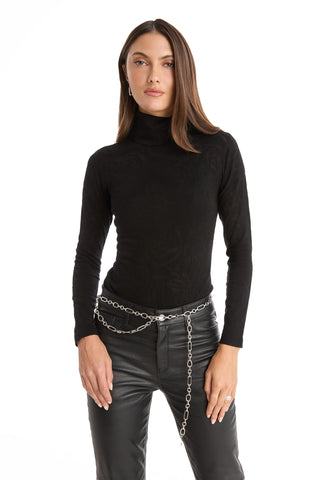 A woman with straight brown hair and a neutral expression stands against a white background wearing the Alina Rose Turtleneck Top in black, paired with black leather pants and a silver chain belt, her hands resting by her sides.