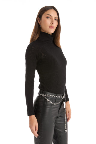 A woman with straight brown hair wears the Alina Rose Turtleneck Top in black, paired with black leather pants and a silver rose-motif chain belt. She stands slightly turned to the side against a white background.