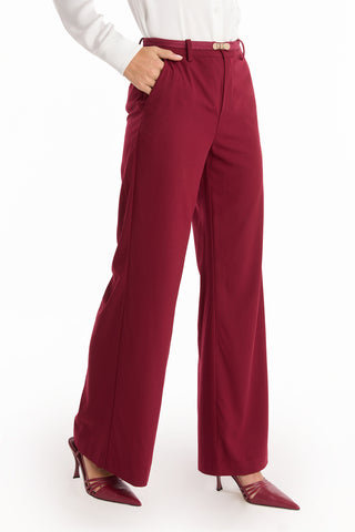 A person stands against a white background, wearing the Martina Belted Trouser in Merlot with a white long-sleeve shirt and maroon pointed-toe heels, one hand in a pocket.