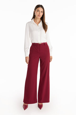 A woman with long brown hair and a neutral expression stands against a white background wearing Martina Belted Trouser - Merlot, paired with a white blouse and matching maroon heels.