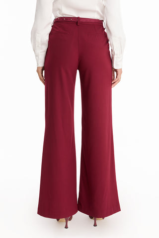 A person is shown from the back wearing the Martina Belted Trouser in Merlot with flare legs and back pockets, styled with a white long-sleeve blouse and heeled shoes, against a white background.