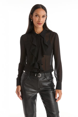 A woman with long, straight dark hair stands facing forward in a Georgia Semi-Sheer Blouse - Black with front ruffles, paired with black leather pants. She has a neutral expression and is set against a white background.