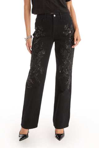 A person wears the Stassi Floral Sequin Jean - Black, featuring intricate black floral embroidery and beading on the front, paired with a black top and pointed-toe heels against a white background.
