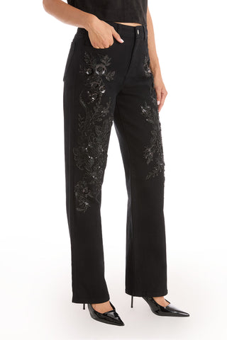 A person wearing the Stassi Floral Sequin Jean - Black, featuring intricate black sequin floral embroidery, paired with black pointed-toe heels, stands against a white background.