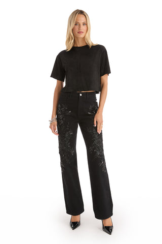 A woman with straight blonde hair wears the Stassi Floral Sequin Jean in black, a black cropped t-shirt, black heels, and bracelets, posing against a white background with a neutral expression.