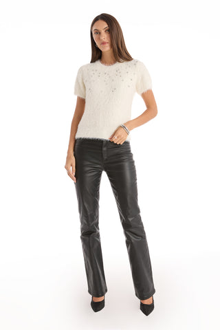 A woman with straight brown hair and a neutral expression poses against a white background wearing the Angel Fluffy Sweater Top - White with embellishments, black leather pants, and black pointed-toe heels.
